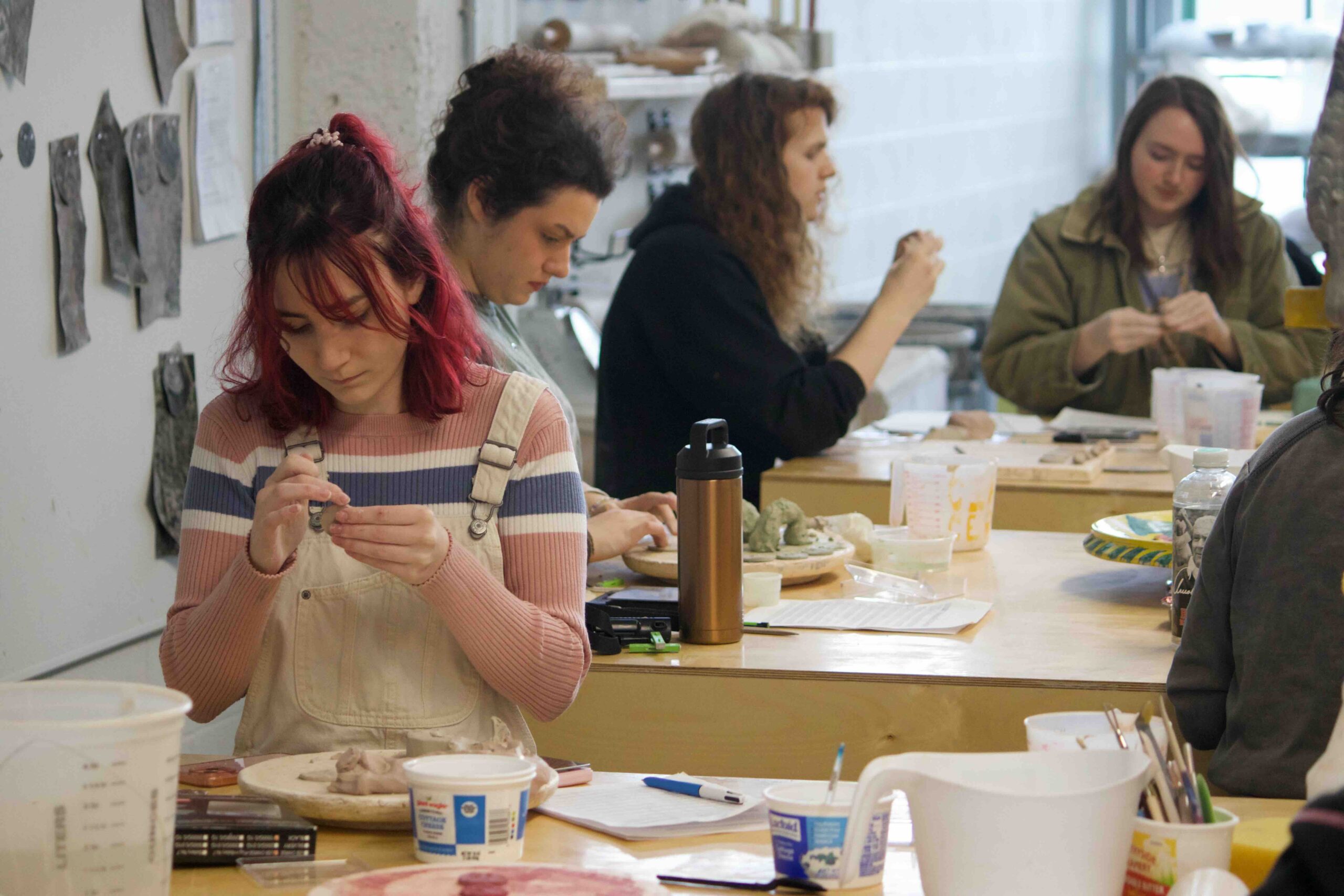 A student is using her hands to work in clay in the ceramics studio. In the background are more students doing the same out of focus.